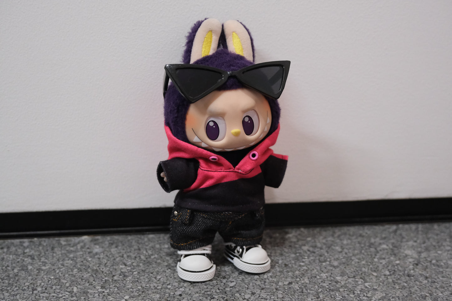 A Labubu figure from the side, wearing a pink and black hoodie that shows the color blocking detail,sunglasses, paired with dark denim shorts.