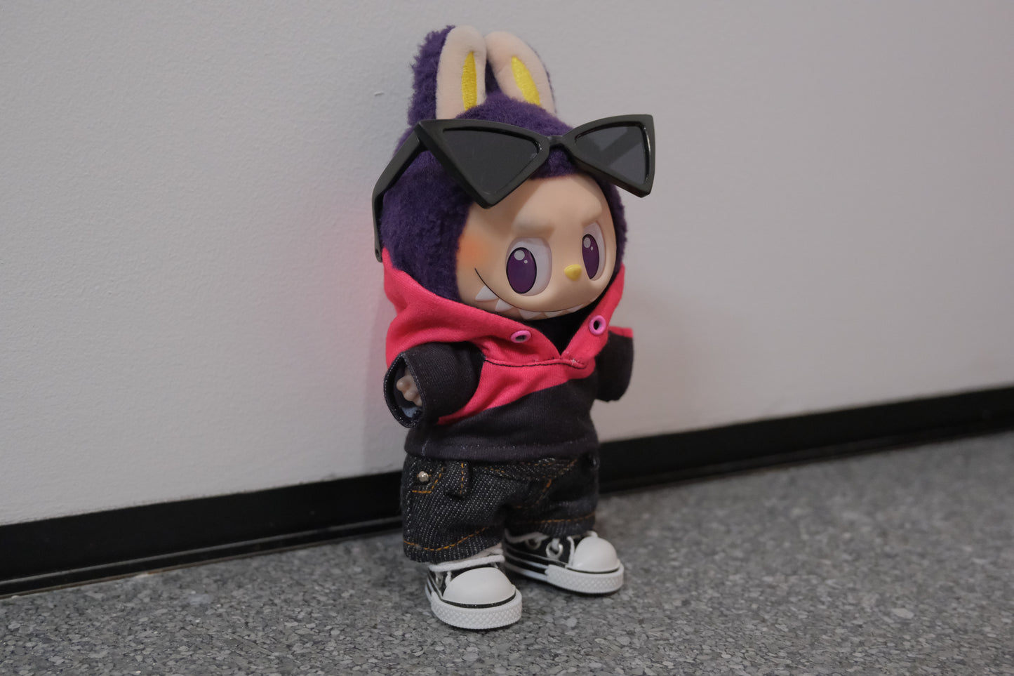 A Labubu figure from the side, wearing a pink and black hoodie that shows the color blocking detail, paired with dark denim shorts.
