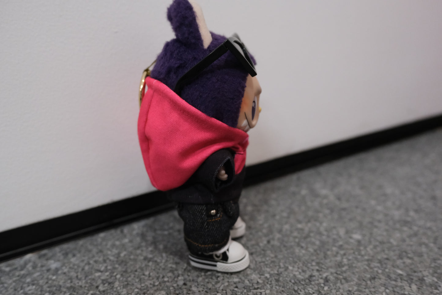 A Labubu figure from the side, wearing a pink and black hoodie that shows the color blocking detail, paired with dark denim shorts.
