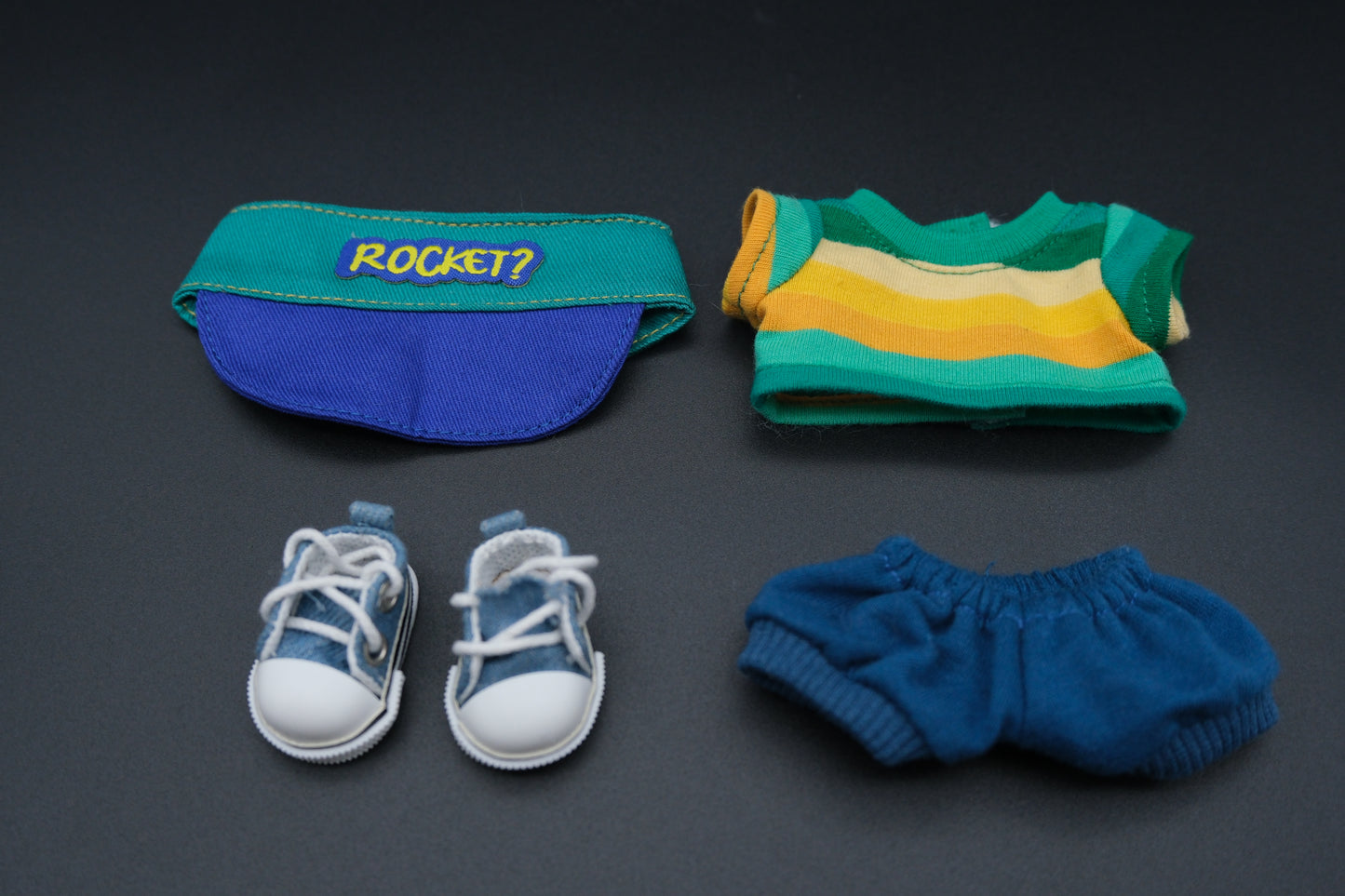 A whimsical Labubu designer toy figure sporting a teal and blue baseball-style cap with a royal blue brim and a yellow "ROCKET?" patch on the front. It's paired with a long-sleeved top featuring bold horizontal stripes in teal, yellow, and white. The figure also wears relaxed-fit blue shorts with a drawstring and classic white sneakers with blue accents and black trim.