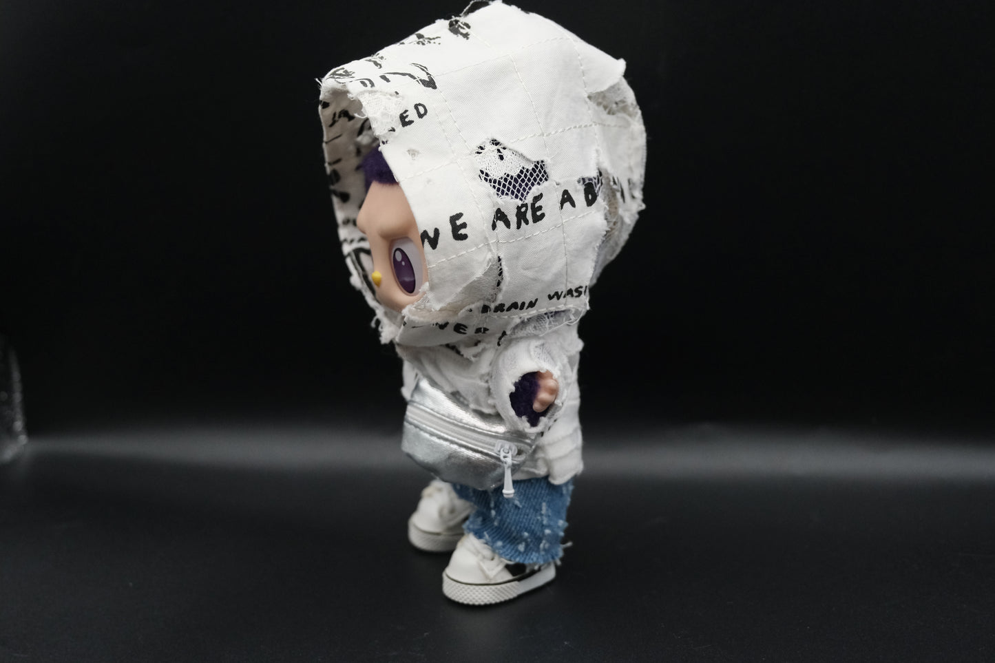A Labubu figure from the side, showcasing the distressed white hooded jacket with black lettering and graphics on the hood, the layered white top, and the distressed blue denim shorts.