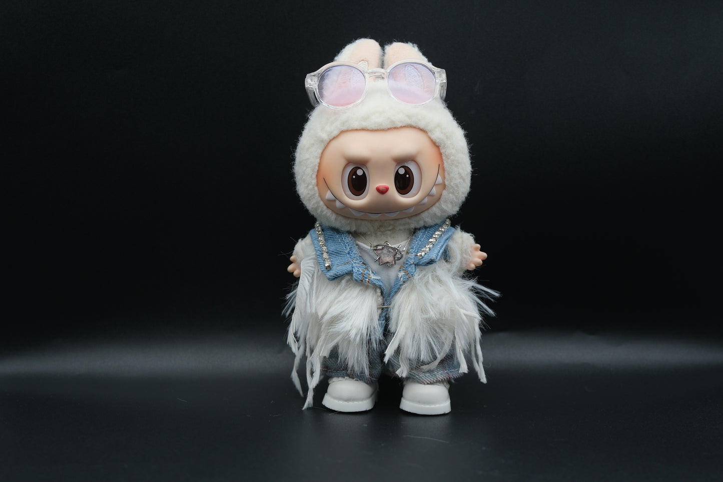 Front view of a Labubu art toy in a unique outfit. The doll wears a light blue denim vest with sparkly rhinestone accents on the shoulders, a white t-shirt, and plaid pants. A white furry, feathery shawl or jacket is layered over the outfit. The doll has sunglasses on its head and a star-shaped necklace.