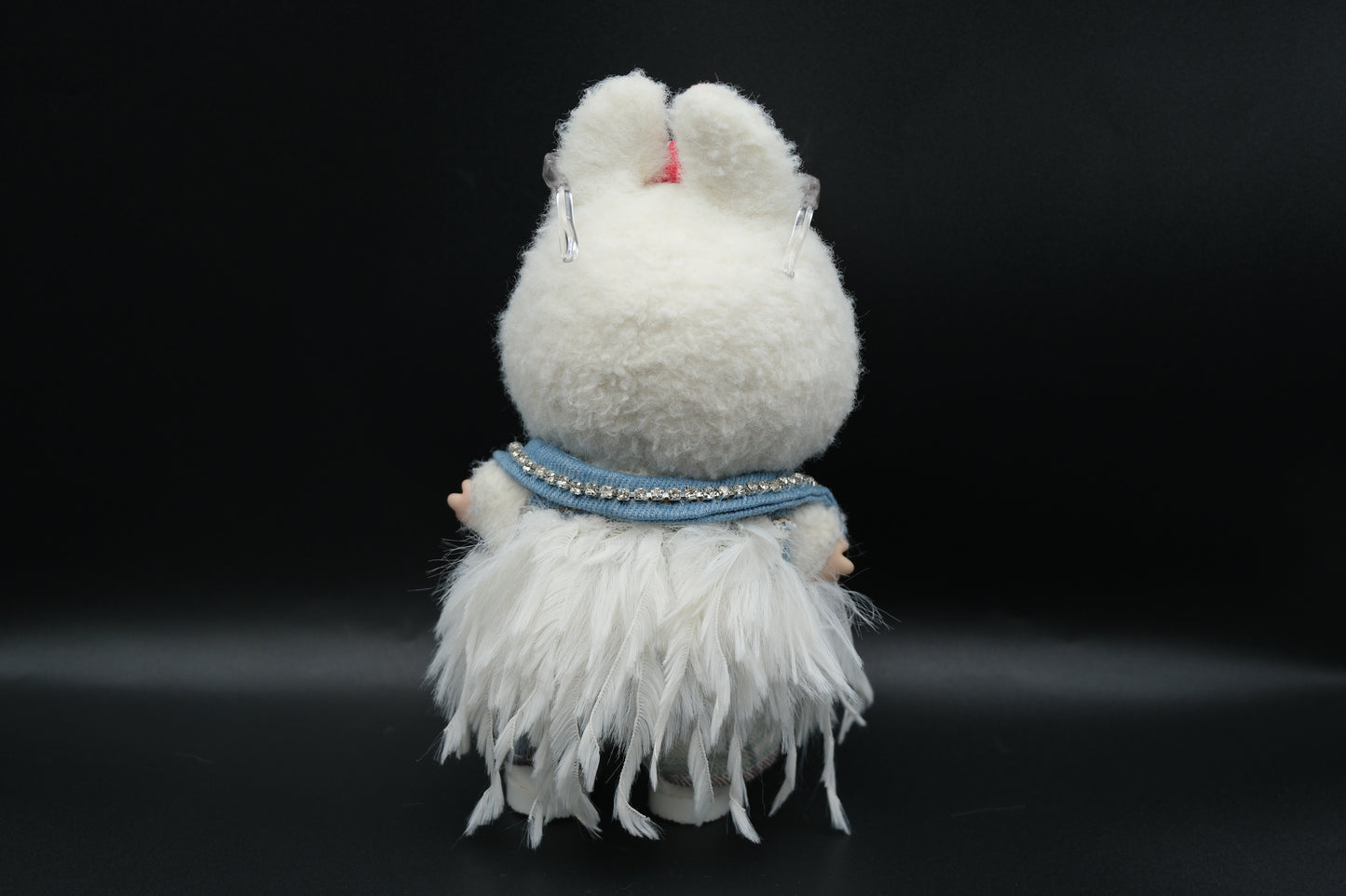 back view of Front view of a Labubu art toy in a unique outfit. The doll wears a light blue denim vest with sparkly rhinestone accents on the shoulders, a white t-shirt, and plaid pants. A white furry, feathery shawl or jacket is layered over the outfit. The doll has sunglasses on its head and a star-shaped necklace.