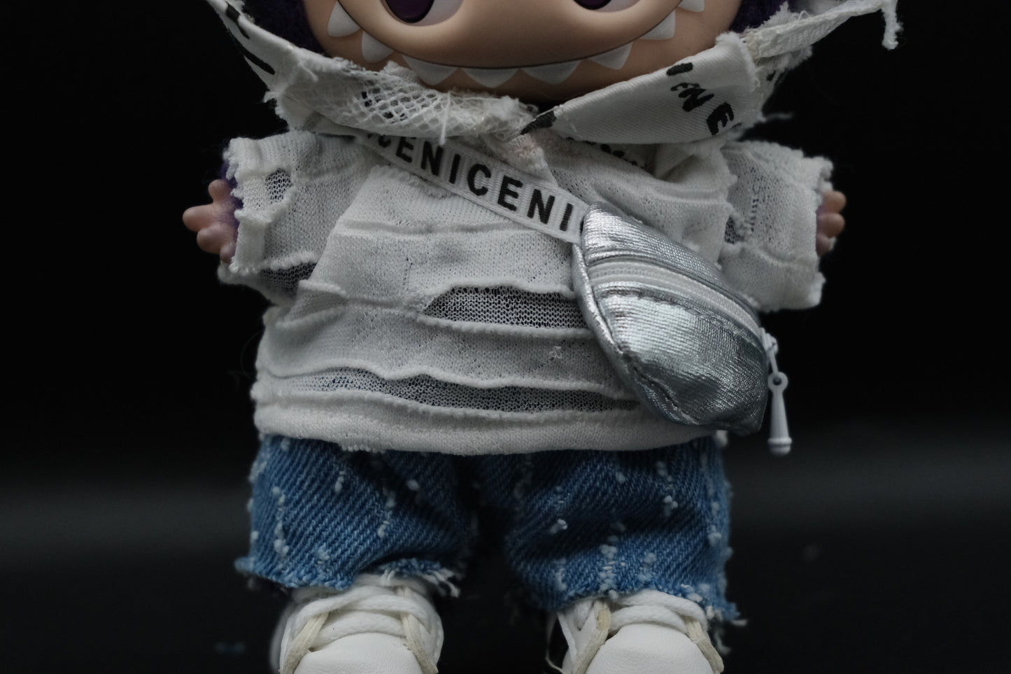 Close-up shot of a Labubu figure showing the layered distressed white graphic hoodie over a ripped white knit top, and a silver crossbody bag with a "ENICENI" branded white strap.