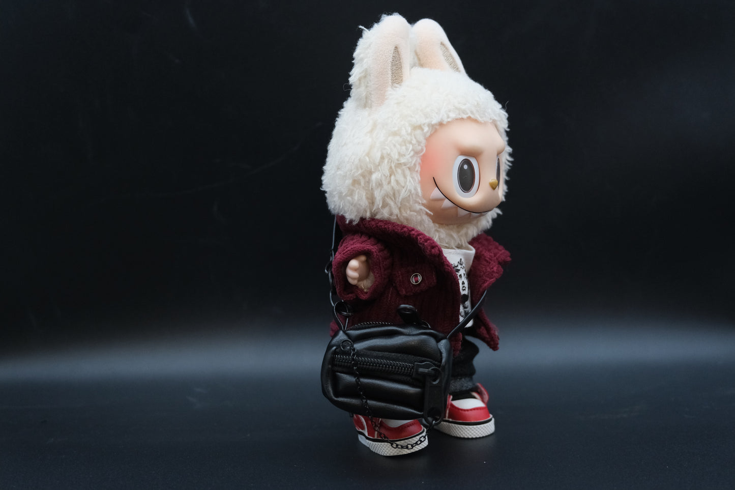 A Labubu figure from the side, wearing a studded nylon jacket, crossbody bag, and showing the details of the t-shirt.