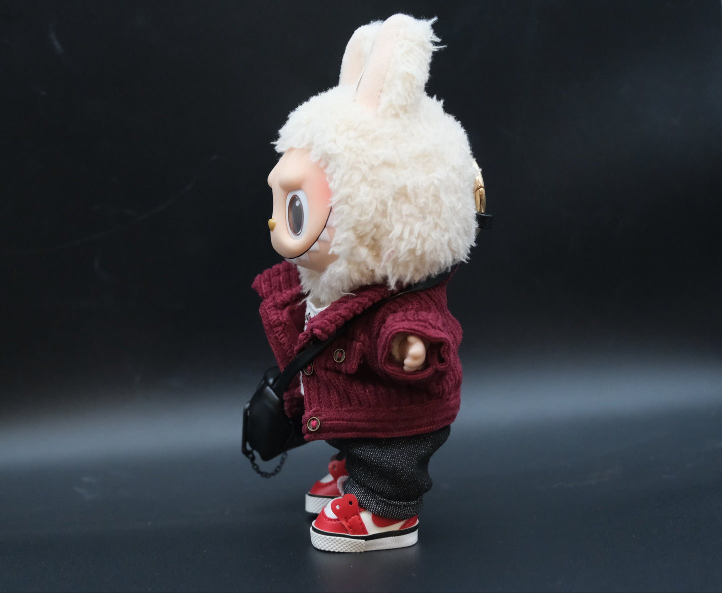 A Labubu figure from the side, wearing a studded nylon jacket, crossbody bag, and showing the details of the t-shirt.