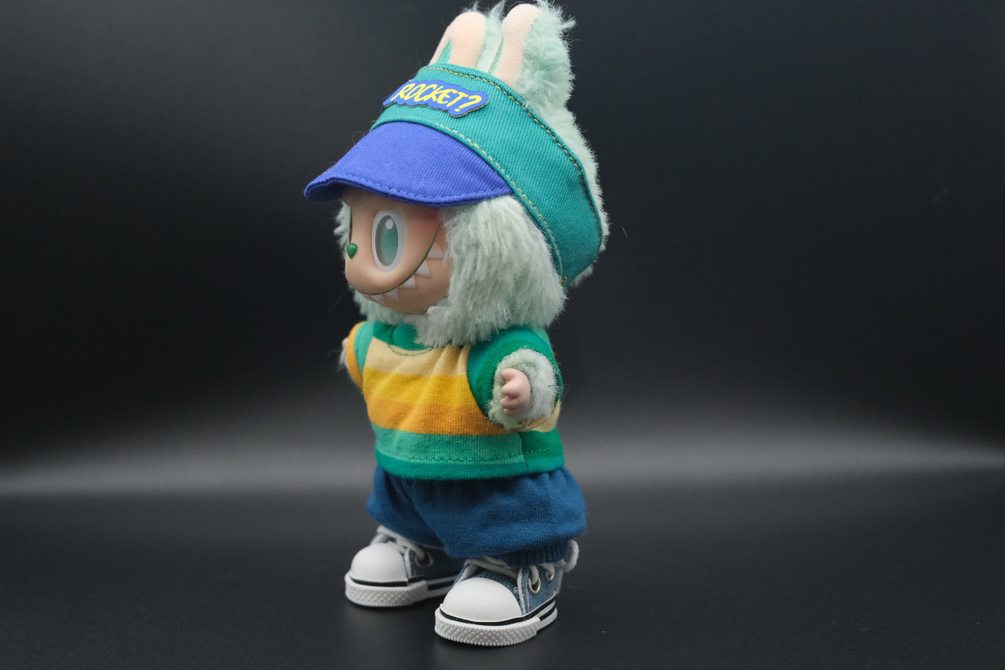 A side view of whimsical Labubu designer toy figure sporting a teal and blue baseball-style cap with a royal blue brim and a yellow "ROCKET?" patch on the front. It's paired with a long-sleeved top featuring bold horizontal stripes in teal, yellow, and white. The figure also wears relaxed-fit blue shorts with a drawstring and classic white sneakers with blue accents and black trim.
