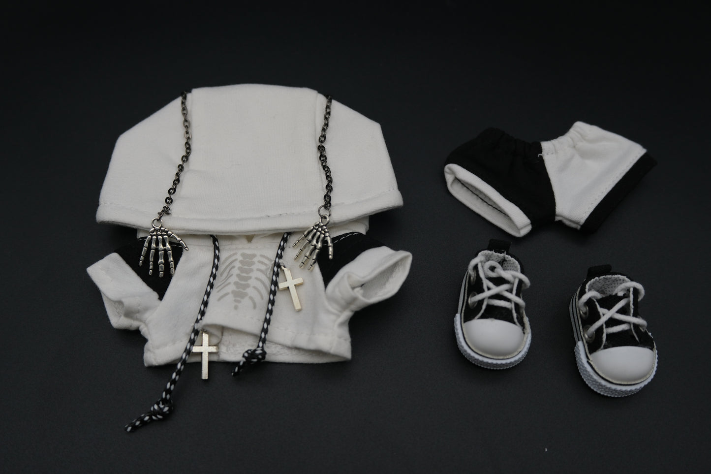 A laid-out set of clothing for a doll or art toy. It includes a white hoodie with a skeleton ribcage graphic, a pair of black and white shorts, and a pair of white high-top sneakers. The hoodie is adorned with two silver skeleton hand charms on chains and two silver cross necklaces.