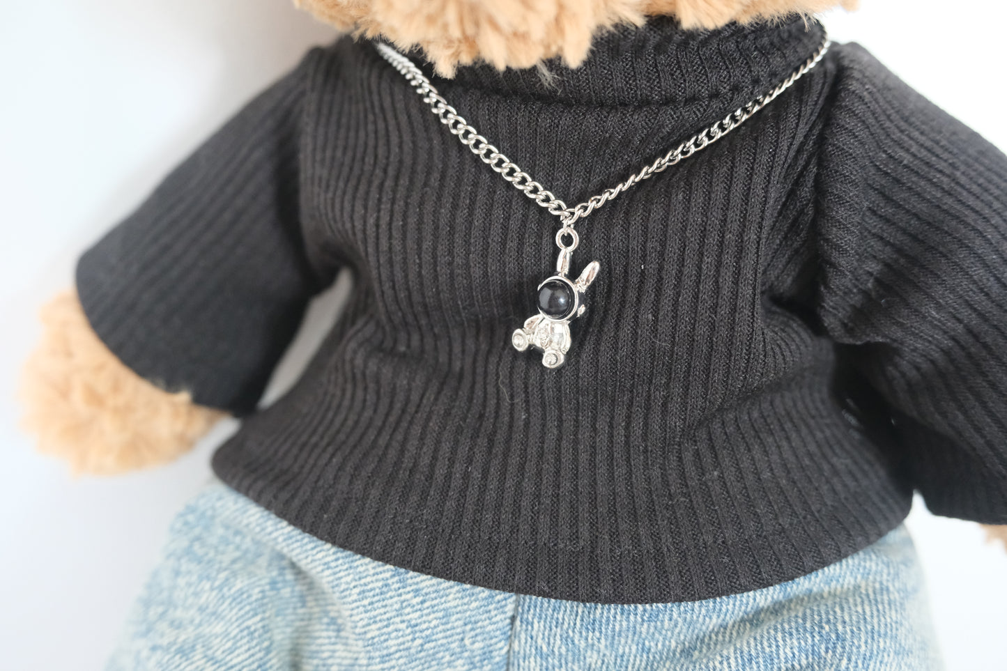 Jellycat bear wearing a dark sweater with a necklace featuring a small pendant.