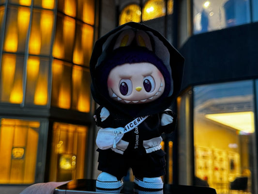 A front-facing, full-body shot of a Labubu art toy. The toy is wearing a black hoodie with white striped sleeves and a white fanny pack with a strap that reads "keep moving" and "nicenice". The doll is also wearing black pants and white sneakers. 
