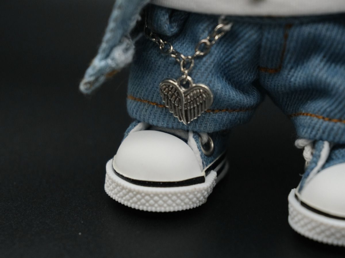 A close-up image showing the details of the doll's denim jeans and a silver heart with wings charm hanging from a chain. The doll's white high-top sneakers are also visible.
