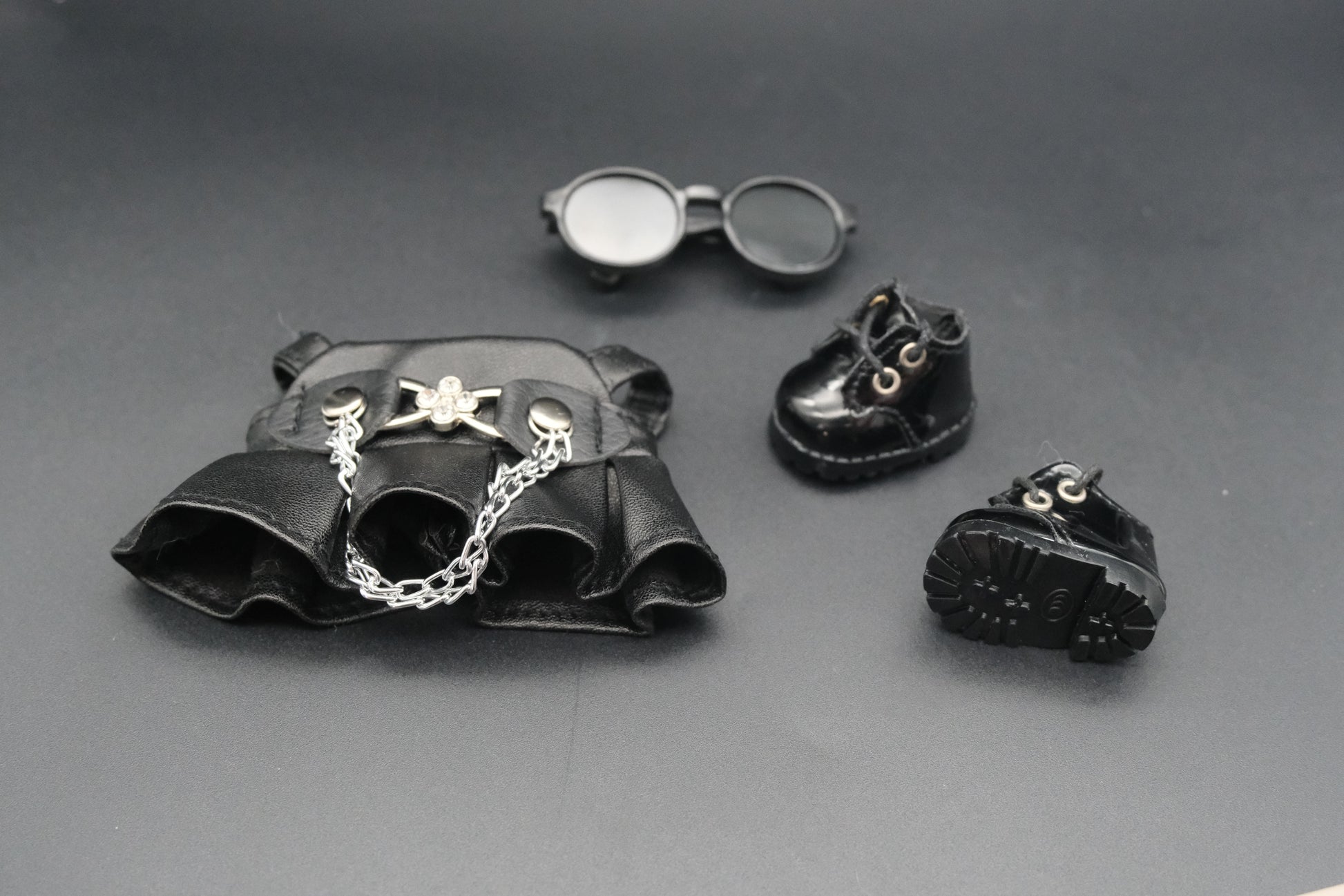 A charming Labubu designer toy figure sporting a chic black faux leather overall dress. The dress features a decorative silver chain detail that adds a touch of edgy flair. Black sunglasses are perched on its head, completing the stylish look.