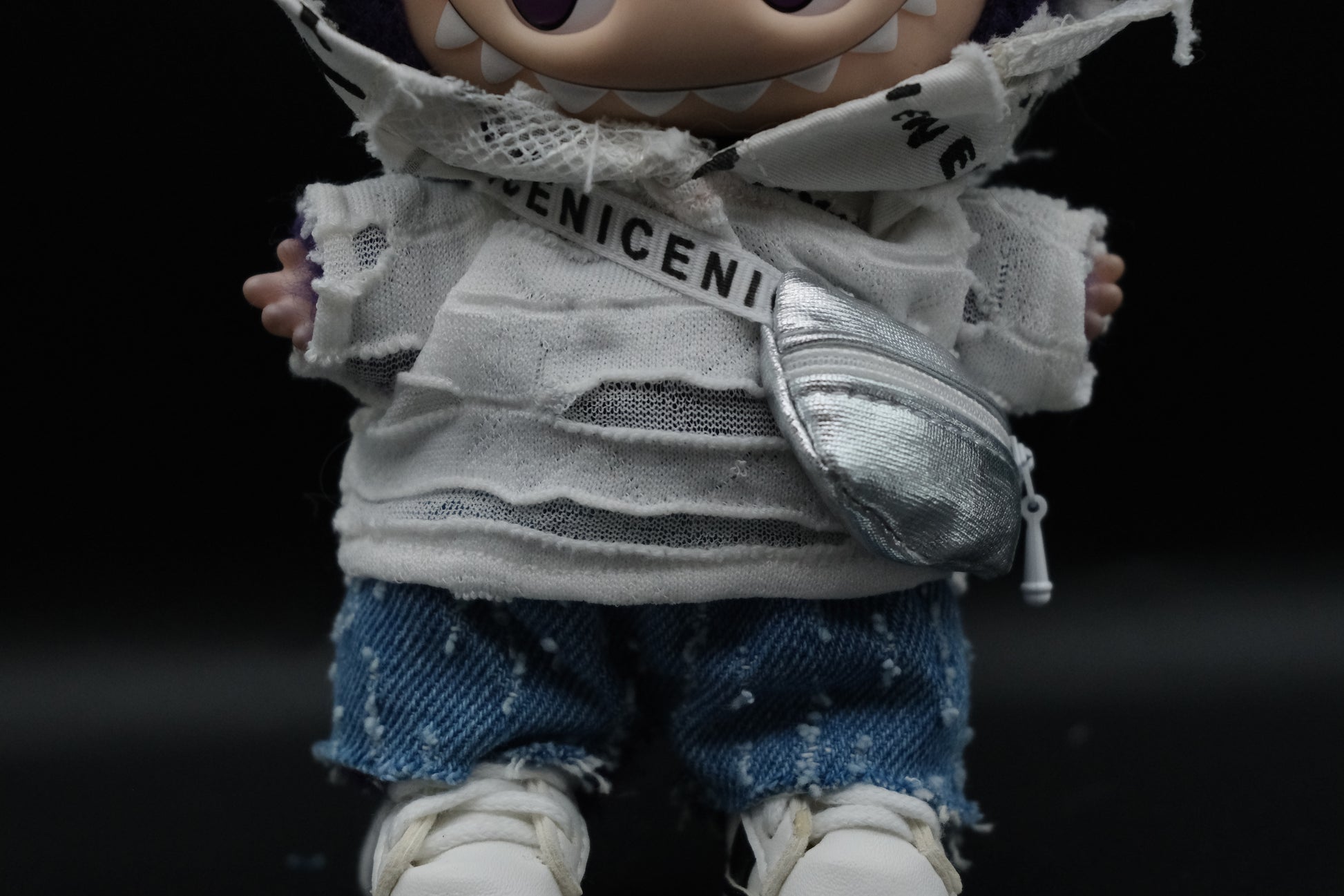 Close-up shot of a Labubu figure showing the layered distressed white graphic hoodie over a ripped white knit top, and a silver crossbody bag with a "ENICENI" branded white strap.