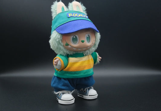A whimsical Labubu designer toy figure sporting a teal and blue baseball-style cap with a royal blue brim and a yellow "ROCKET?" patch on the front. It's paired with a long-sleeved top featuring bold horizontal stripes in teal, yellow, and white. The figure also wears relaxed-fit blue shorts with a drawstring and classic white sneakers with blue accents and black trim.