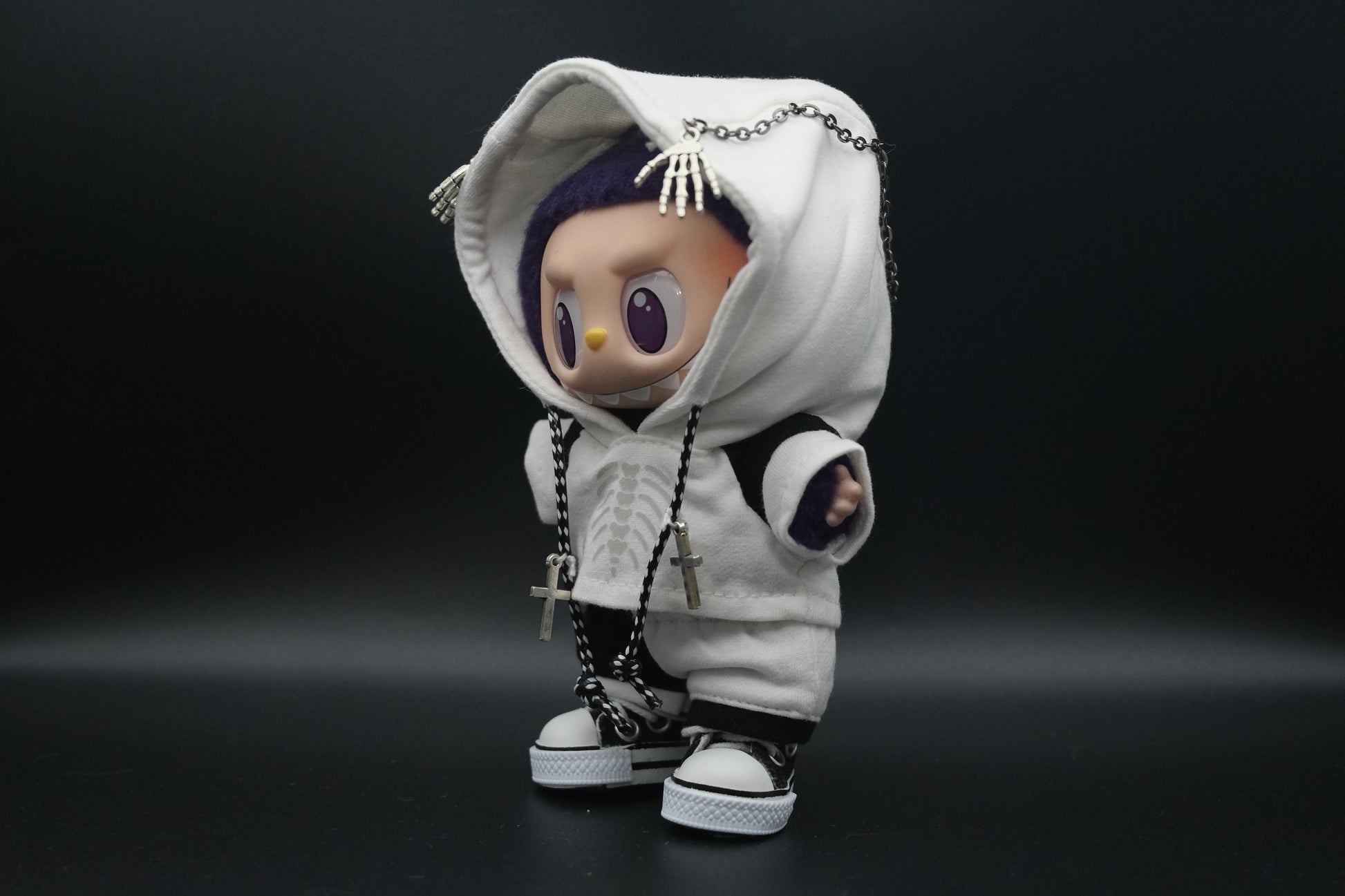 An angled, full-body shot of a Labubu doll wearing a white hoodie with a skeleton ribcage graphic, a skeleton hand chain, and black and white shorts. The doll's fuzzy purple head and yellow-lined bunny ears are visible.