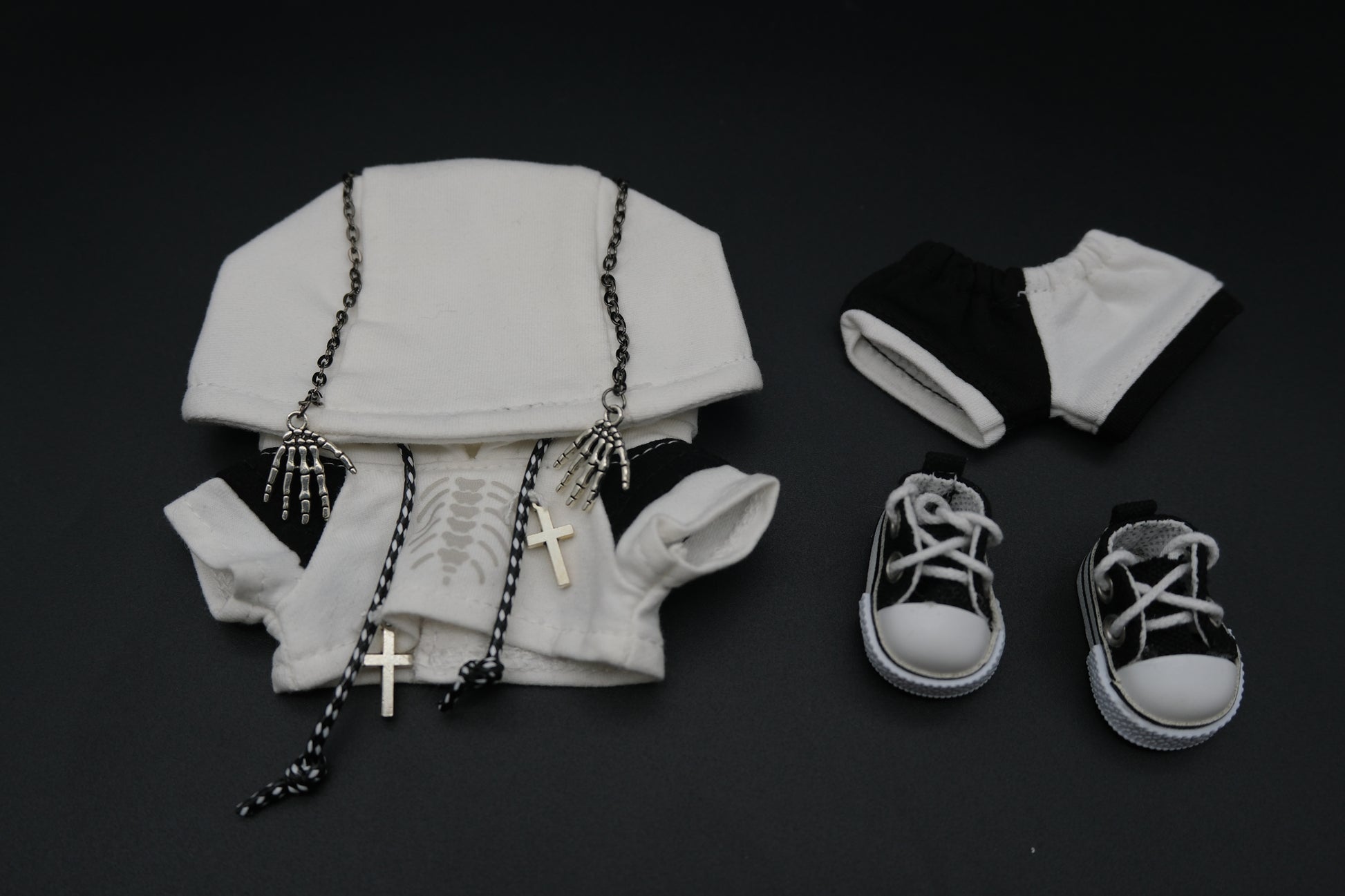 A laid-out set of clothing for a doll or art toy. It includes a white hoodie with a skeleton ribcage graphic, a pair of black and white shorts, and a pair of white high-top sneakers. The hoodie is adorned with two silver skeleton hand charms on chains and two silver cross necklaces.
