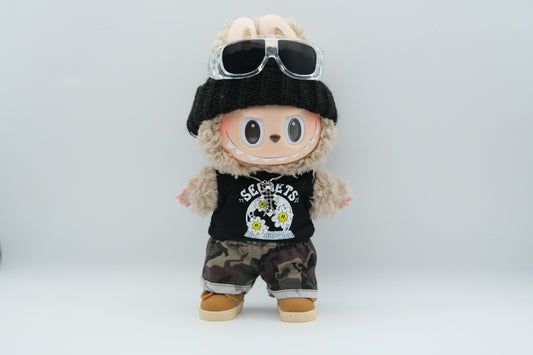A Labubu figure wearing a black knit beanie, a black tank top with a white "SECRETS" and floral graphic, camouflage print shorts with rolled cuffs, and tan sneakers with white soles. A silver cross pendant necklace and sunglasses are also visible.