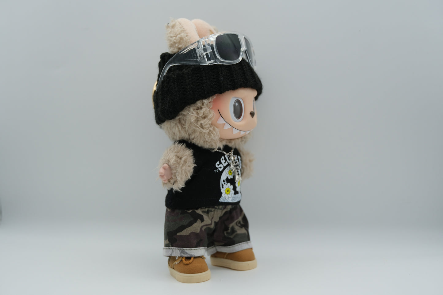 side view of A Labubu figure wearing a black knit beanie, a black tank top with a white "SECRETS" and floral graphic, camouflage print shorts with rolled cuffs, and tan sneakers with white soles. A silver cross pendant necklace and sunglasses are also visible.