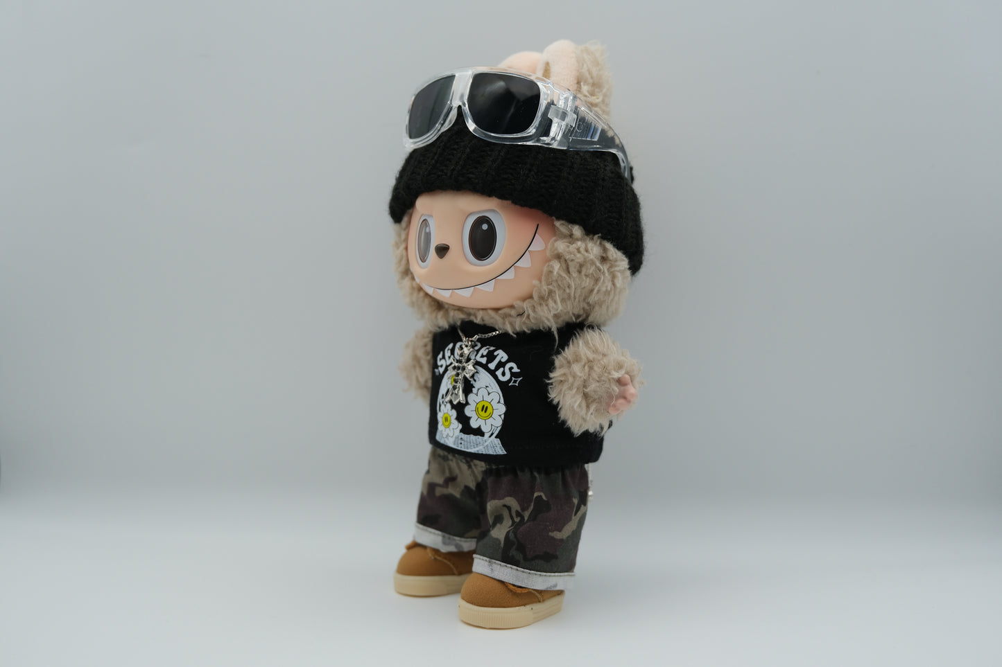 side view of A Labubu figure wearing a black knit beanie, a black tank top with a white "SECRETS" and floral graphic, camouflage print shorts with rolled cuffs, and tan sneakers with white soles. A silver cross pendant necklace and sunglasses are also visible.