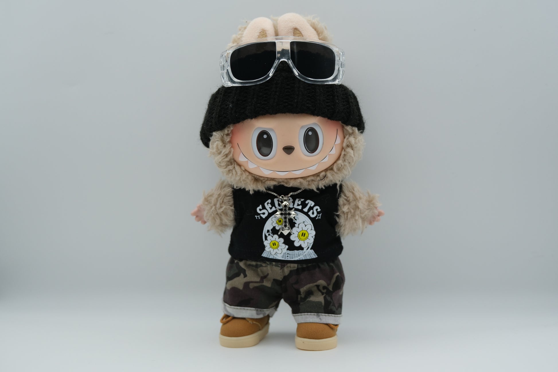 A Labubu figure wearing a black knit beanie, a black tank top with a white "SECRETS" and floral graphic, camouflage print shorts with rolled cuffs, and tan sneakers with white soles. A silver cross pendant necklace and sunglasses are also visible.