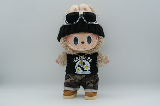 A Labubu figure wearing a black knit beanie, a black tank top with a white "SECRETS" and floral graphic, camouflage print shorts with rolled cuffs, and tan sneakers with white soles. A silver cross pendant necklace and sunglasses are also visible.