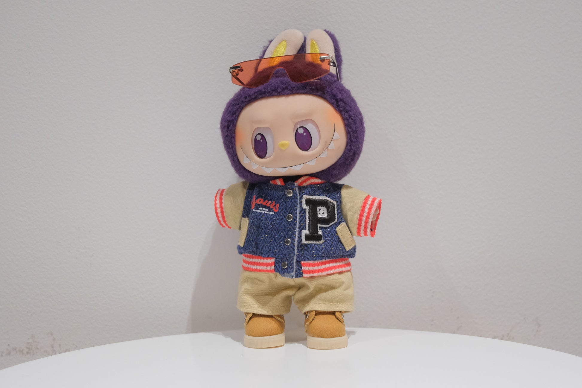 Labubu figure wearing a 4-piece varsity streetwear outfit with a baseball jacket (P-patch), sporty sunglasses, khaki shorts, and boots.