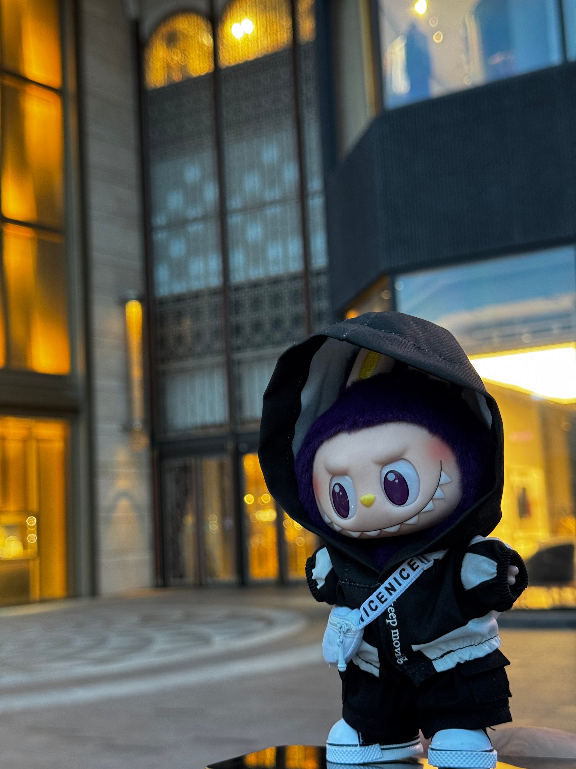 A front-facing, full-body shot of a Labubu art toy. The toy is wearing a black hoodie with white striped sleeves and a white fanny pack with a strap that reads "keep moving" and "nicenice". The doll is also wearing black pants and white sneakers. 