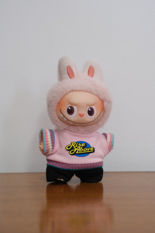 pin for love Labubu figure wearing a light pink sweater with a "Rise Above" graphic and black pants