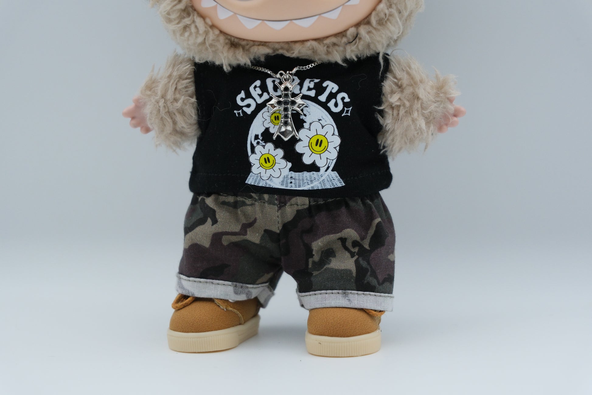 details of A Labubu figure wearing  a black tank top with a white "SECRETS" and floral graphic, camouflage print shorts with rolled cuffs, and tan sneakers with white soles. A silver cross pendant necklace and sunglasses are also visible.