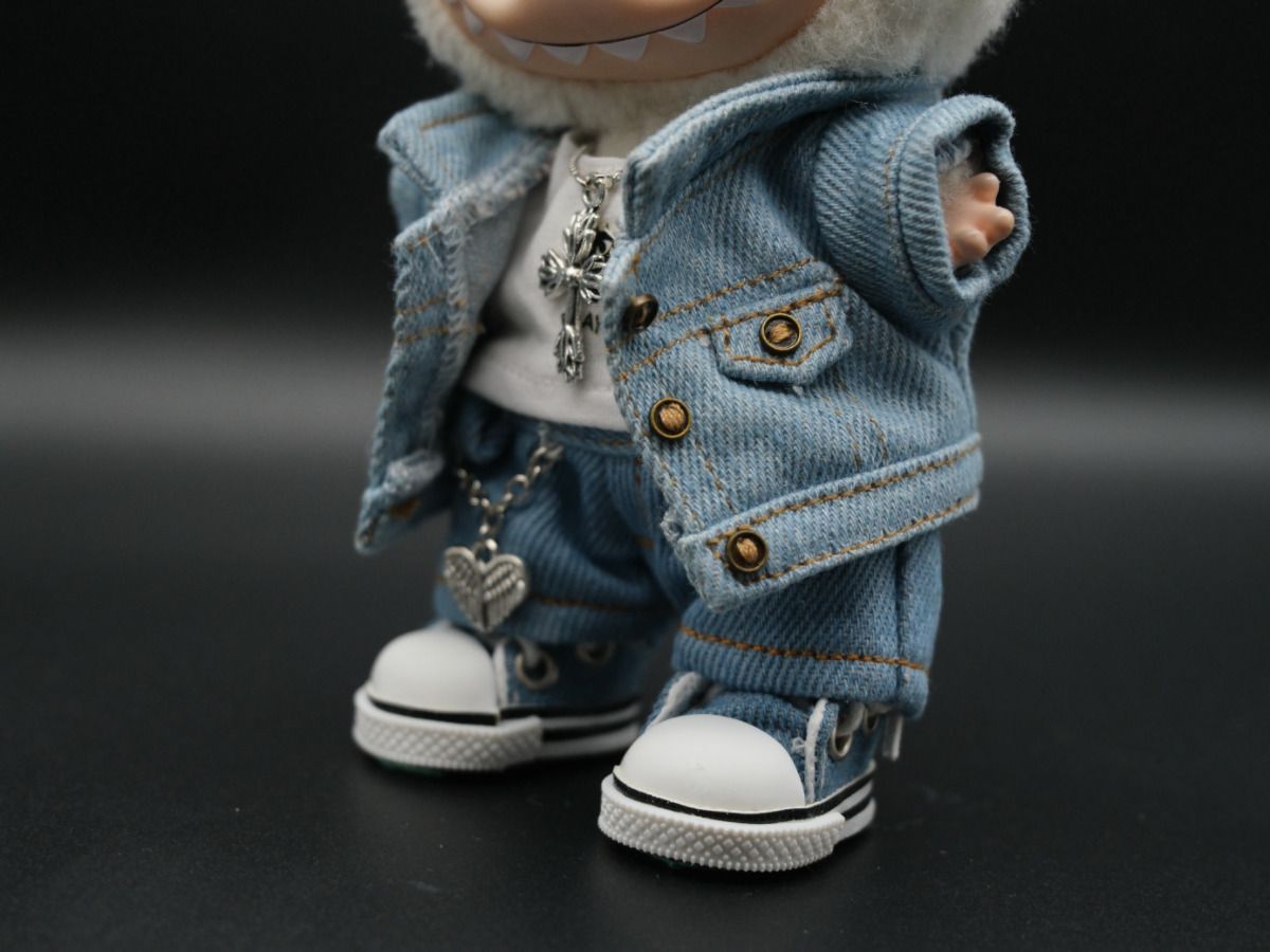A close-up image showing the details of the doll's denim jeans and a silver heart with wings charm hanging from a chain. The doll's white high-top sneakers are also visible.

