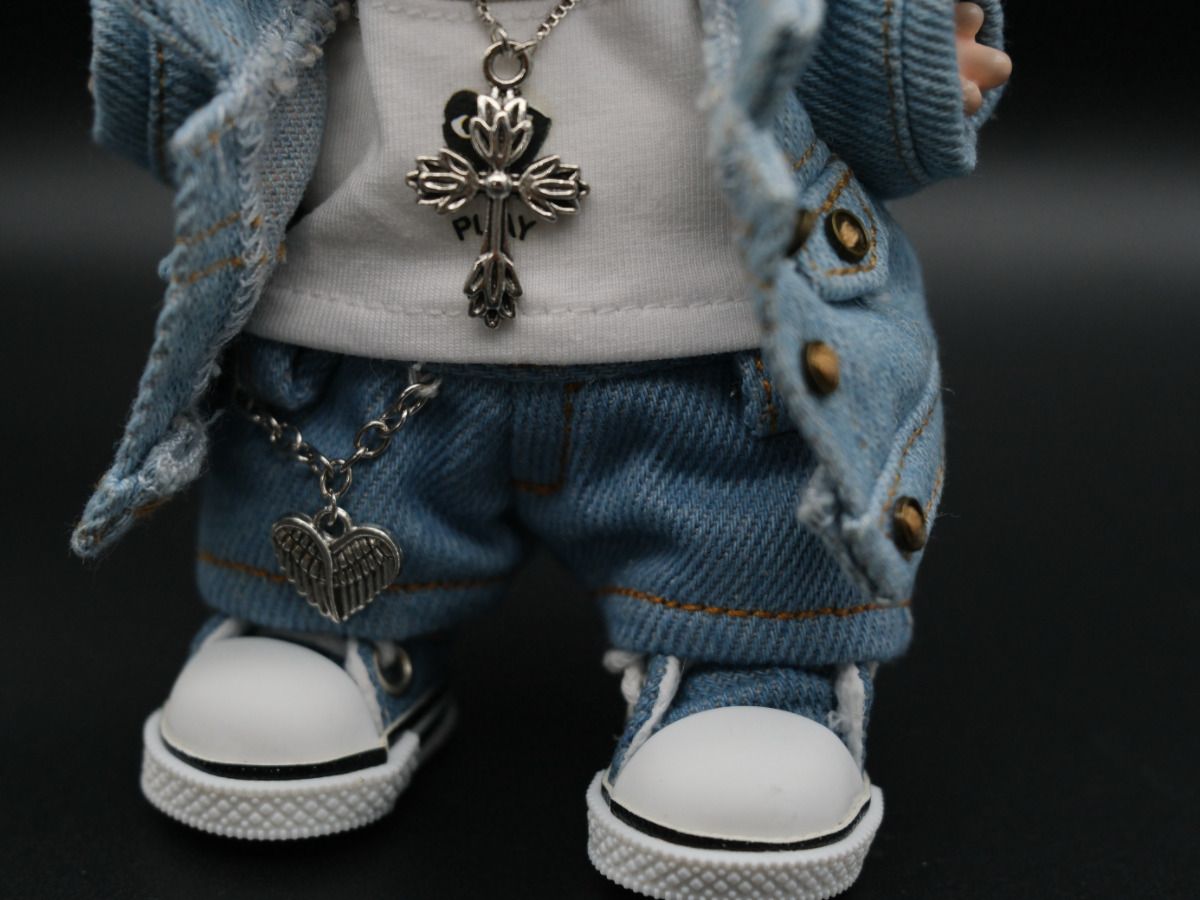 A close-up image showing the details of the doll's denim jeans and a silver heart with wings charm hanging from a chain. The doll's white high-top sneakers are also visible.
